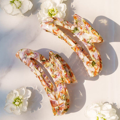 Tia Clip van Clips, een elegante haarclip in een kleurrijke marmerafwerking, perfect voor fijn of glad haar, met een achtergrond van bloemetjes.